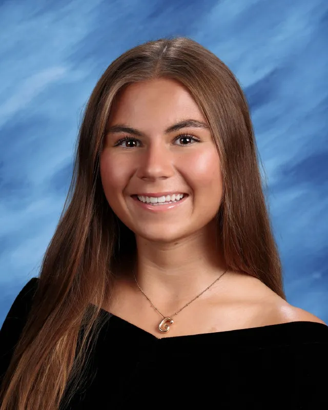 Smiling young woman with long brown hair wearing a black off-shoulder top and a gold necklace against a blue studio background.