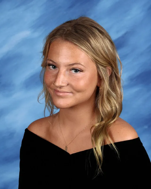 Smiling young woman with long blonde hair, wearing a black off-the-shoulder top against a blue studio background.