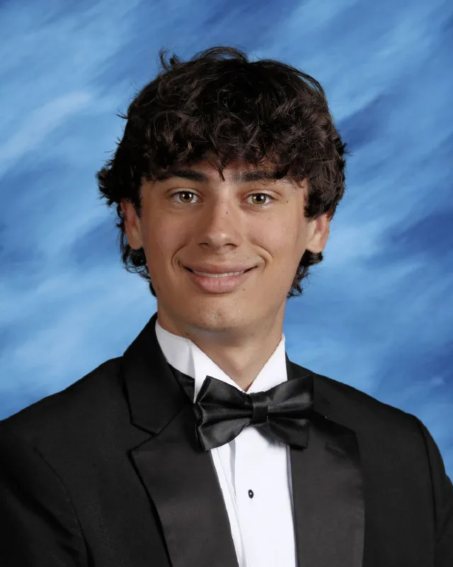 Young man with curly dark hair smiling, wearing a black tuxedo and bow tie against a blue background.