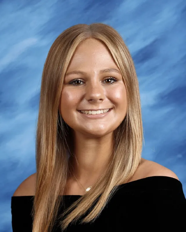 Portrait of a young woman with straight blonde hair wearing a black off-shoulder top and a necklace, smiling against a blue textured background.