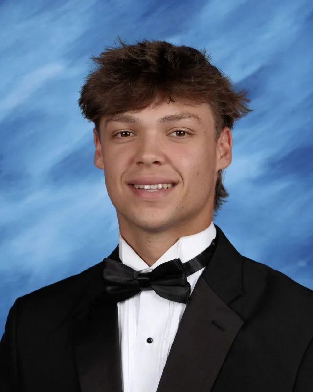 Young man with brown hair wearing a black tuxedo, white shirt, and black bow tie against a blue background.