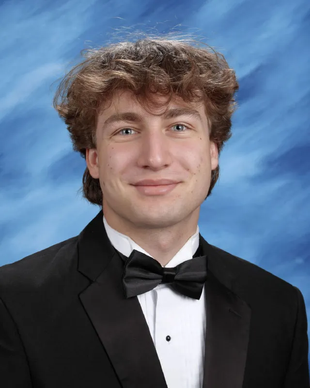 Young man with curly brown hair wearing a black tuxedo jacket, white dress shirt, and black bow tie against a blue cloudy background.