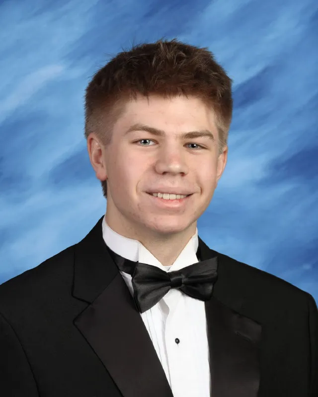 Young man smiling, dressed in a black tuxedo with a white shirt and black bow tie against a blue studio backdrop.