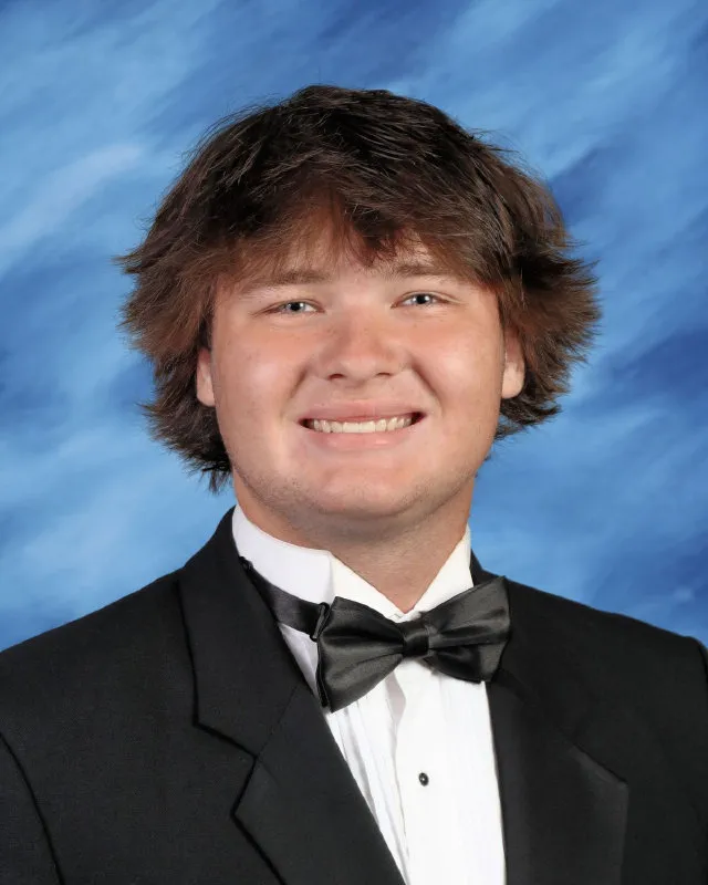 Young man with medium-length brown hair wearing a black tuxedo and bow tie, smiling against a blue backdrop.