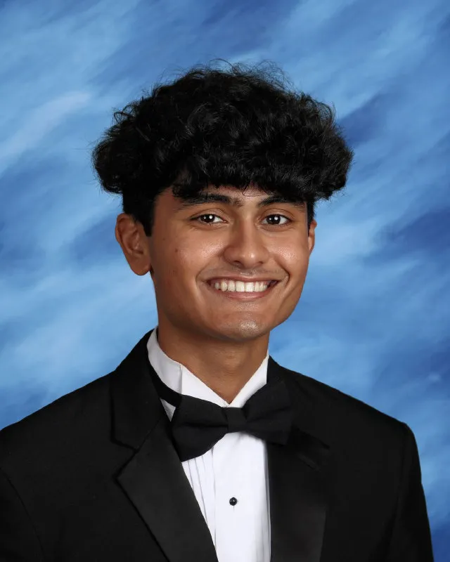 Young man with curly hair wearing a black tuxedo and bow tie smiling against a blue backdrop.