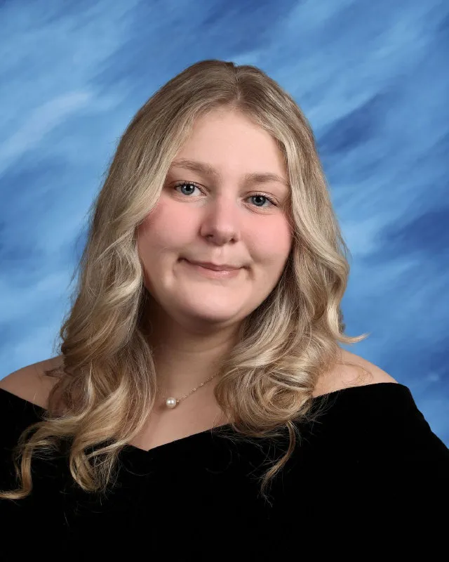 Young woman with long wavy blonde hair wearing a black off-shoulder top and pearl necklace against a blue background.