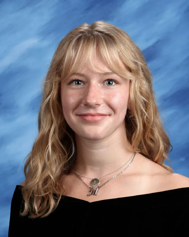 Blonde young woman with wavy hair wearing a black off-shoulder top and two silver necklaces, smiling against a blue marbled background.