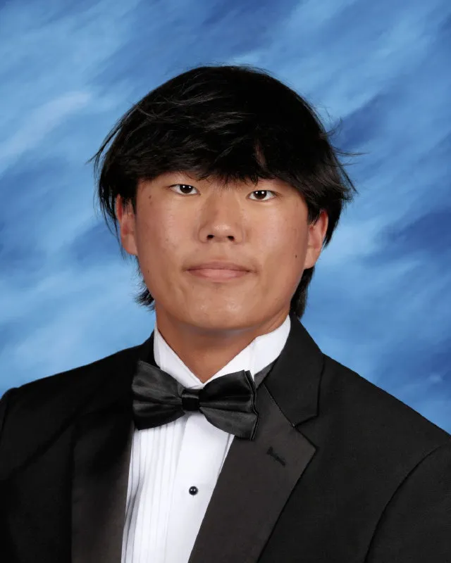 Young man with black hair wearing a black tuxedo, white shirt, and black bow tie against a blue cloudy background.