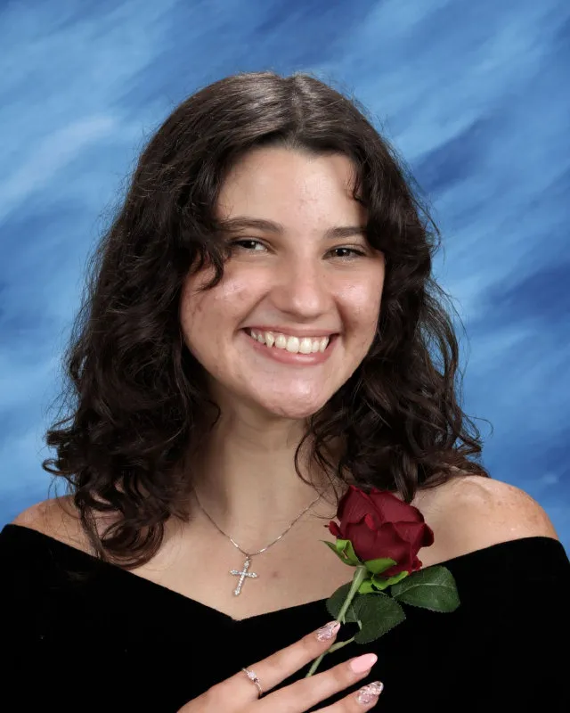 Smiling young woman with curly dark hair holding a red rose against a blue backdrop, wearing a black off-shoulder top and a cross necklace.