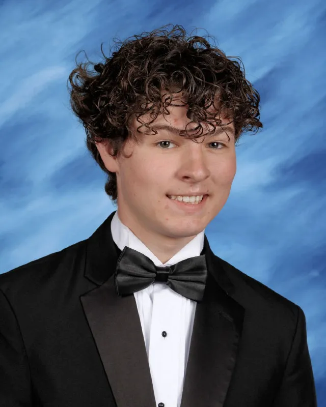 Young man with curly hair wearing a black tuxedo and bow tie against a blue backdrop.