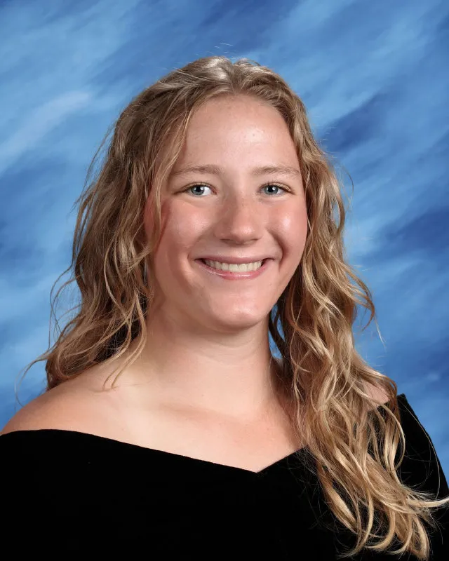 Smiling young woman with wavy blonde hair wearing a black off-shoulder top against a blue backdrop.