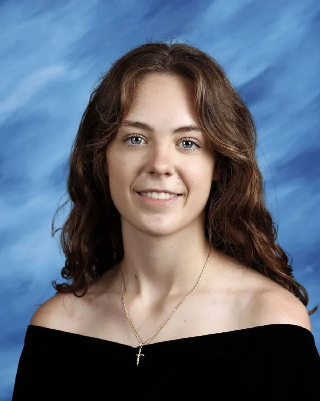 Young woman with long wavy brown hair wearing a black off-shoulder top and a gold cross necklace, smiling against a blue backdrop.