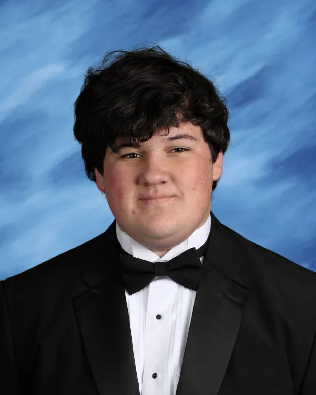 Portrait of a young man with dark hair wearing a black tuxedo and bow tie against a blue backdrop.