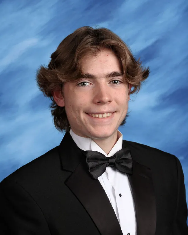 Young man with medium-length brown hair wearing a black tuxedo, white shirt, and black bow tie, smiling against a blue background.