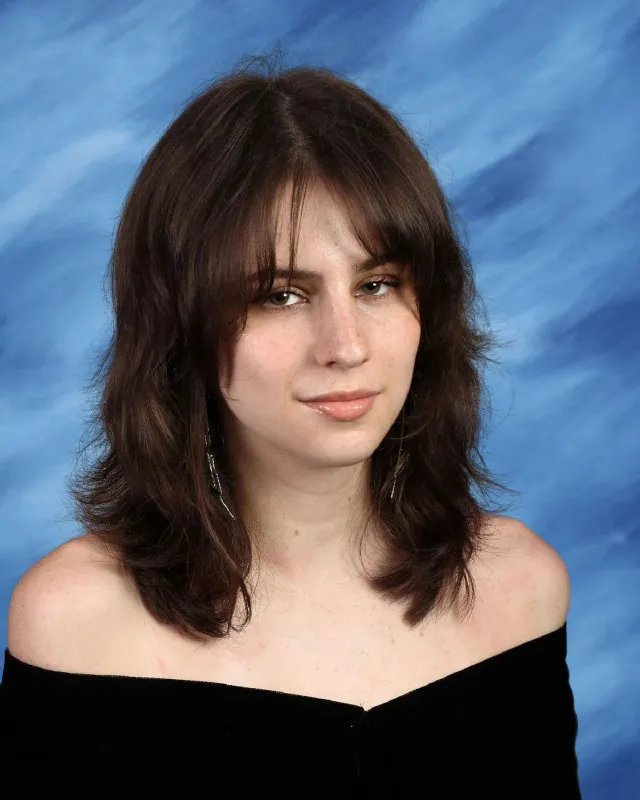 Young woman with medium-length brown hair wearing a black off-shoulder top against a blue studio backdrop.