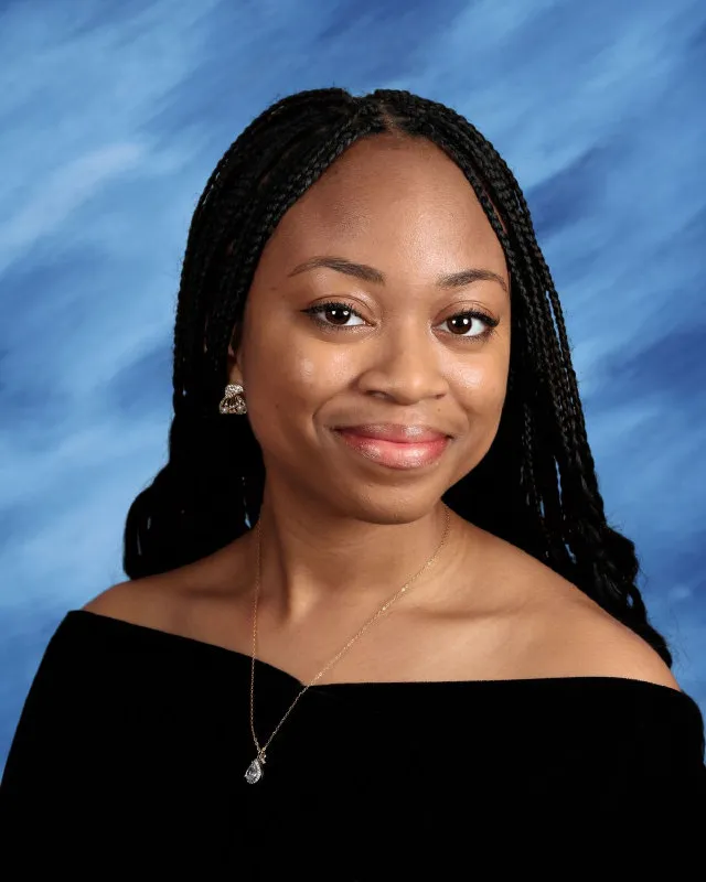 Smiling young woman with braided hair wearing a black off-shoulder top and a necklace against a blue background.