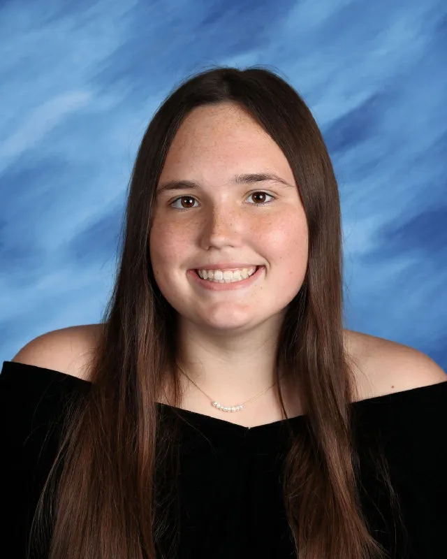 Smiling young woman with long brown hair wearing an off-the-shoulder black top and a delicate necklace with pearls, against a blue cloud-like background.