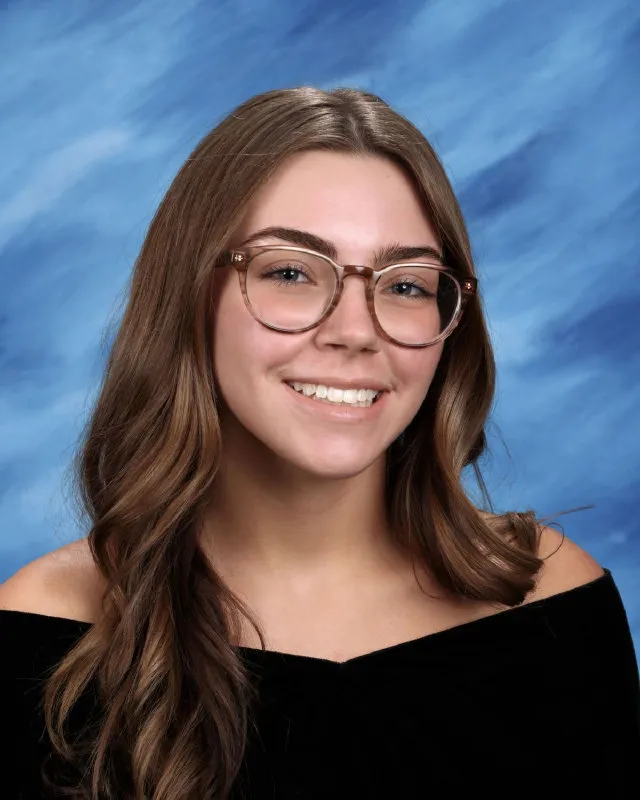 Smiling young woman with long wavy brown hair and glasses, wearing a black off-shoulder top against a blue gradient background.