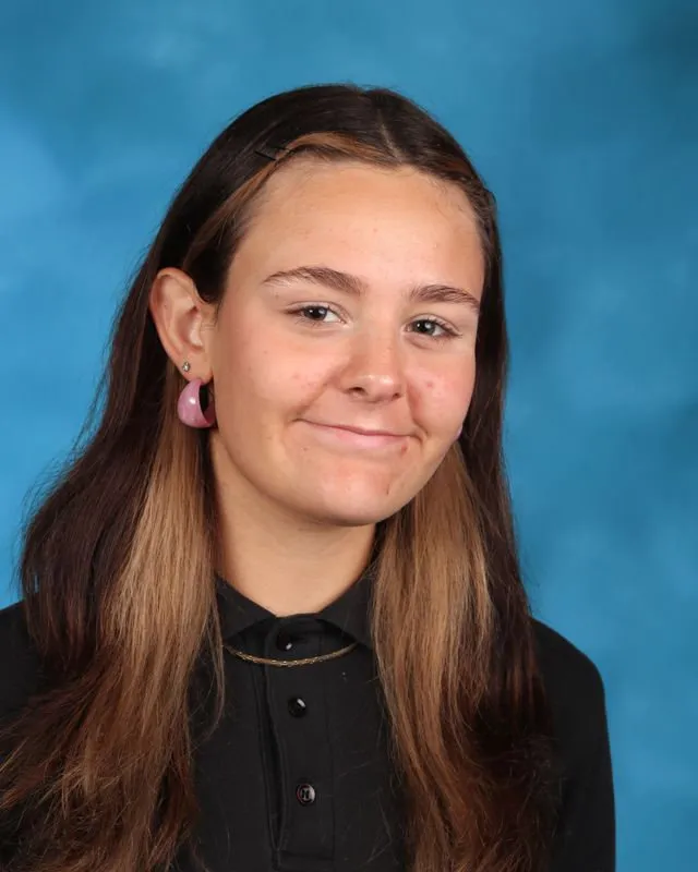 Portrait of a teenage girl with long two-toned brown hair wearing pink hoop earrings and a black collared shirt, smiling slightly against a blue background.