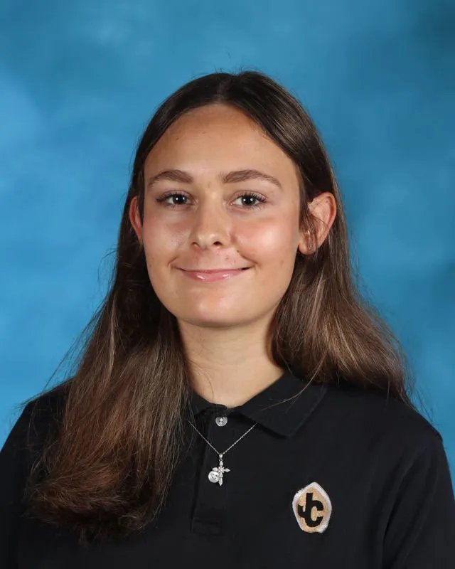 Portrait of a young woman with long brown hair wearing a black collared shirt with a JC logo and a silver necklace, against a blue background.