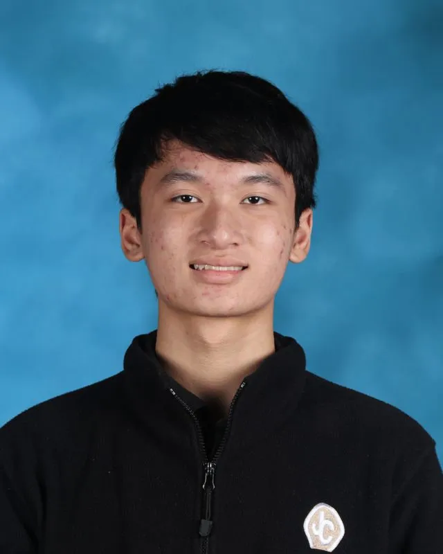 Young man with short black hair wearing a black zip-up jacket against a blue background.
