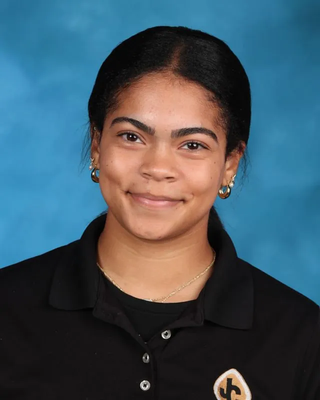 Young woman with dark hair, wearing a black collared shirt with a logo on it, smiling against a blue background.