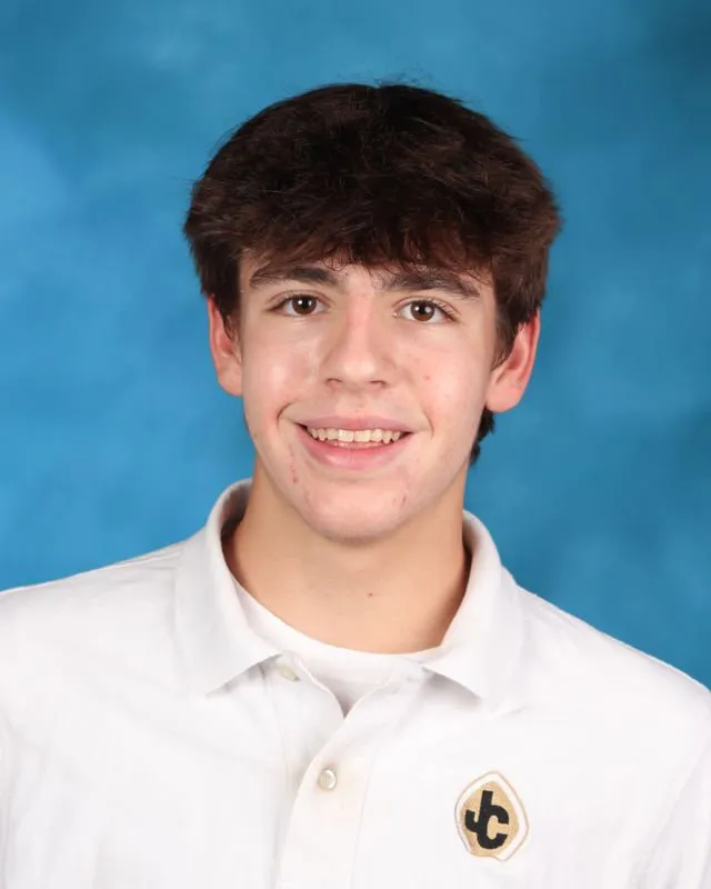 Smiling teenage boy with brown hair wearing a white collared shirt with a JC logo on a blue background.