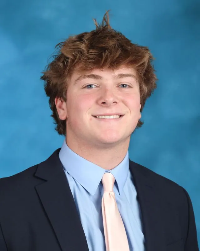 Young man with tousled brown hair wearing a dark blazer, light blue shirt, and pale pink tie smiling against a blue background.