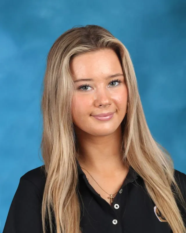 Young woman with long blonde hair wearing a black collared shirt and a cross necklace, against a blue background.