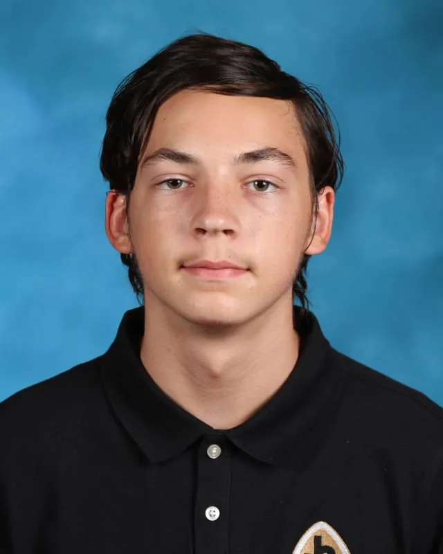 Portrait of a young man with short dark hair wearing a black polo shirt against a blue background.