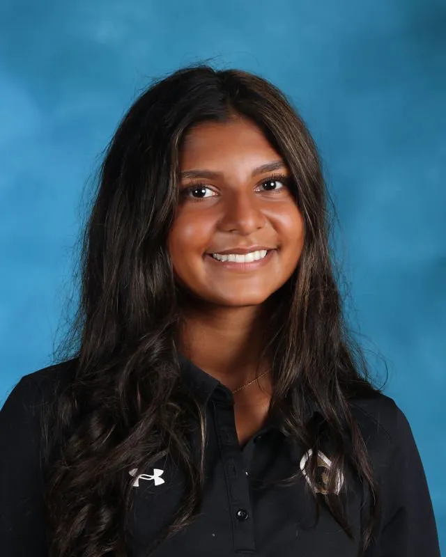 Smiling young woman with long dark hair wearing a black Under Armour polo shirt against a blue background.