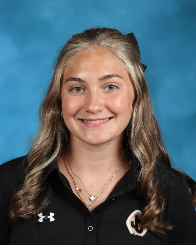 Smiling young woman with long wavy hair wearing a black collared shirt with a sports logo on a blue background.