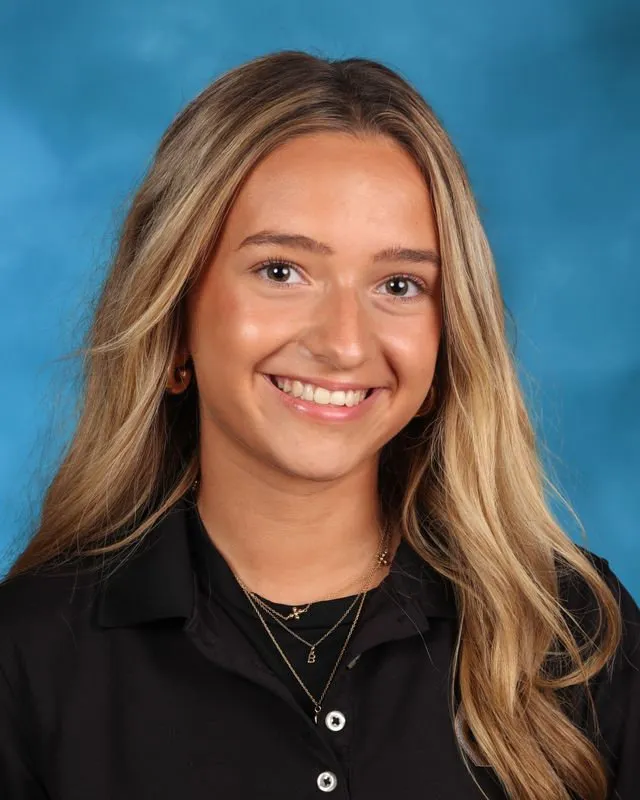 Smiling young woman with long blonde hair wearing a black collared shirt and layered necklaces against a blue background.
