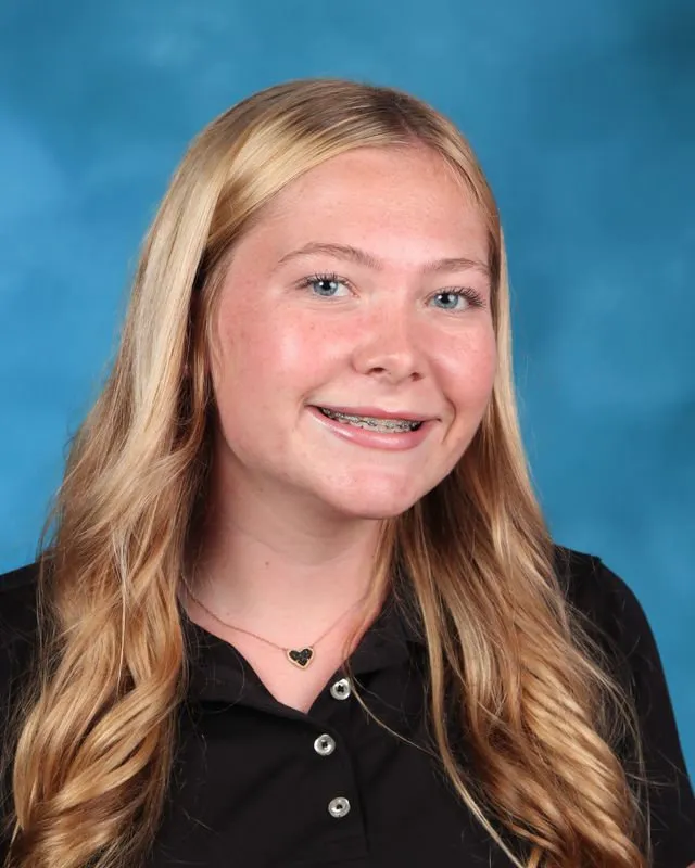 Smiling teenage girl with long blonde hair, wearing a black collared shirt and a heart-shaped necklace, against a blue background.