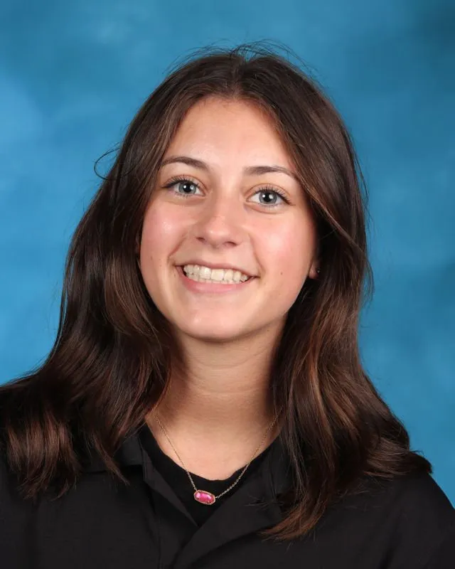Young woman with long brown hair smiling against a blue background, wearing a black top and pink pendant necklace.
