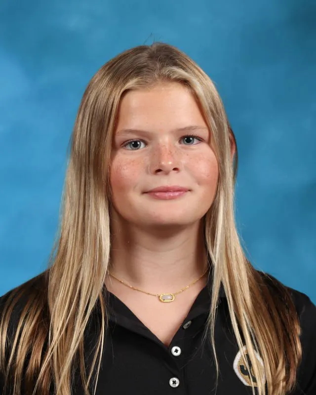 Portrait of a young blonde girl with light freckles, wearing a black collared shirt and a gold necklace, against a blue background.