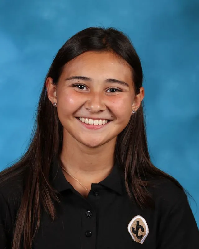 Smiling young woman with long dark hair wearing a black polo shirt with a JC logo patch on a blue background.