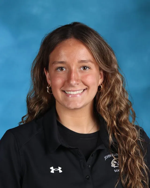 Smiling young woman with wavy brown hair wearing a black John Carroll Soccer polo shirt against a blue background.
