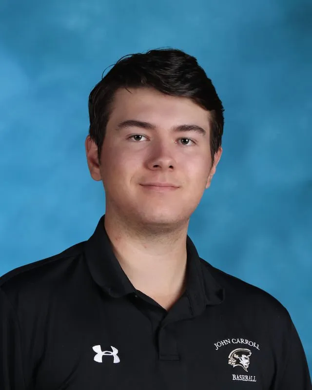 Young man wearing a black John Carroll Baseball polo shirt with a blue studio background.