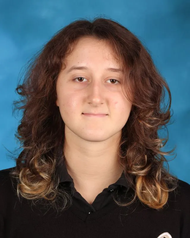 Portrait of a young person with medium-length curly brown hair with blonde tips, wearing a black collared shirt, against a blue background.