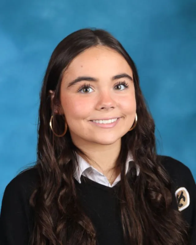 Smiling young woman with long dark hair wearing gold hoop earrings and a black sweater with a patch, against a blue background.