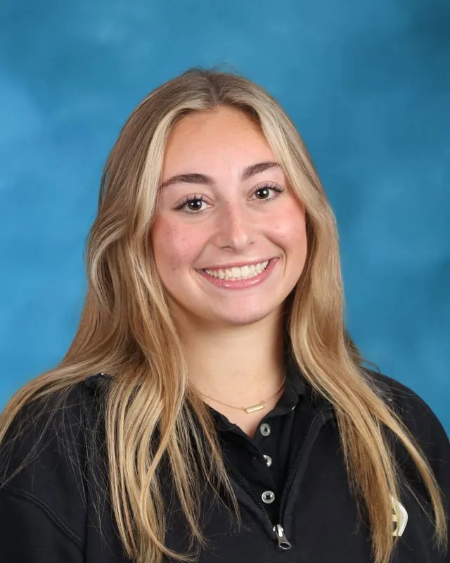 Smiling young woman with long blonde hair wearing a black top against a blue background.