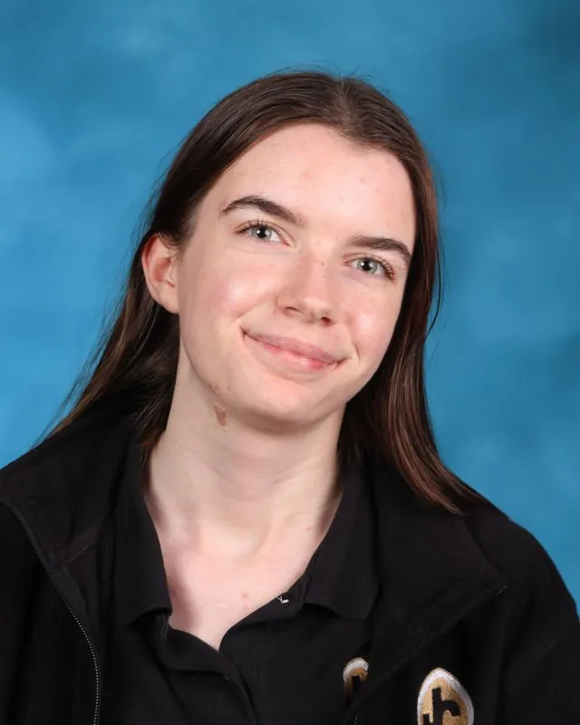 Young woman with long brown hair wearing a black jacket and shirt, smiling against a blue background.