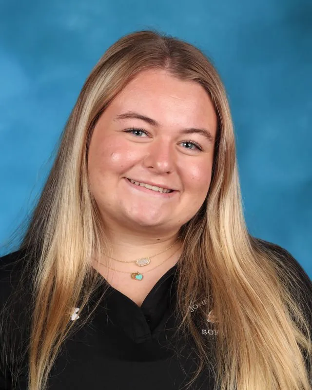 Smiling young woman with long blonde hair wearing a black shirt and layered necklaces against a blue backdrop.