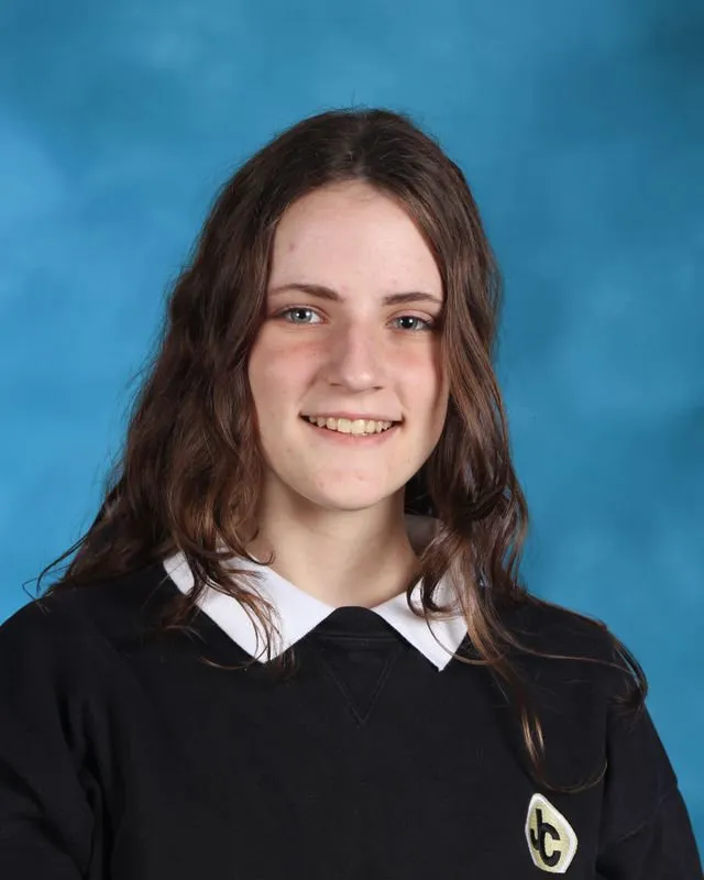 Smiling teenage girl with long wavy brown hair wearing a black top with a white collar against a blue background.