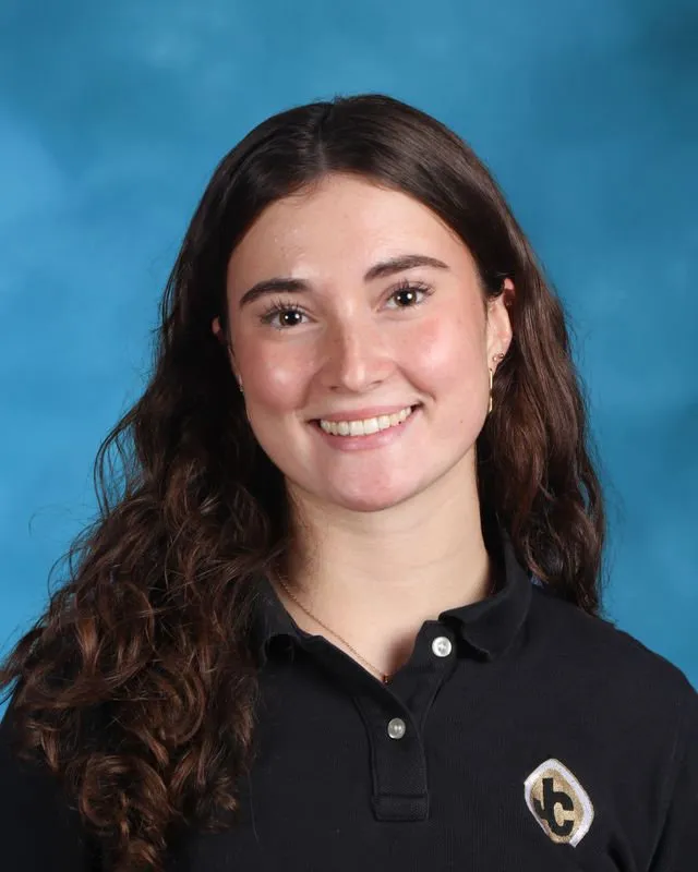 Young woman with long curly brown hair smiling, wearing a black polo shirt with a 'lc' emblem, against a blue background.
