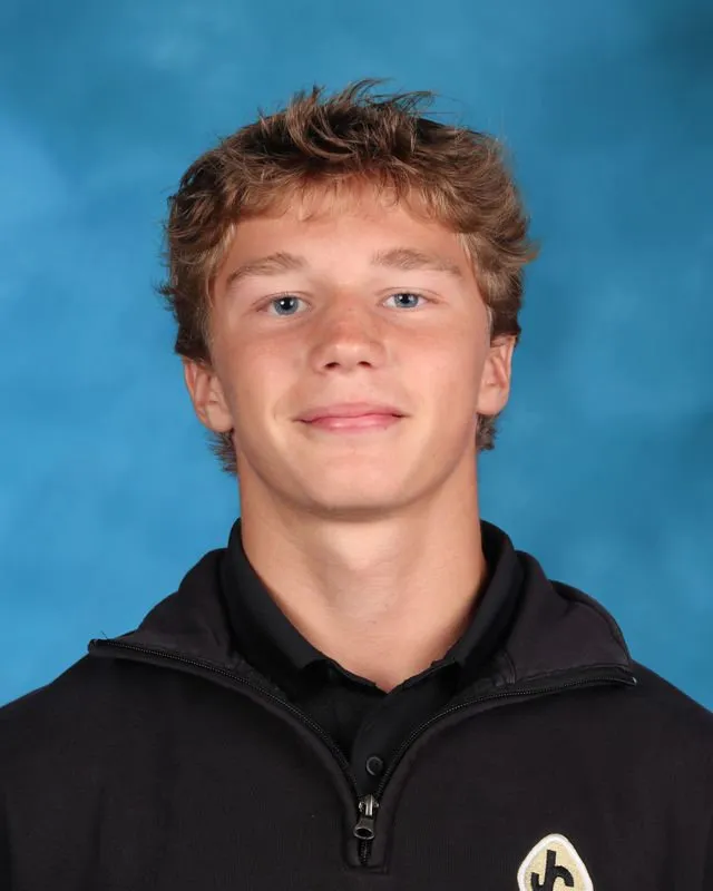 Young man with light brown hair wearing a black zip-up jacket against a blue background.