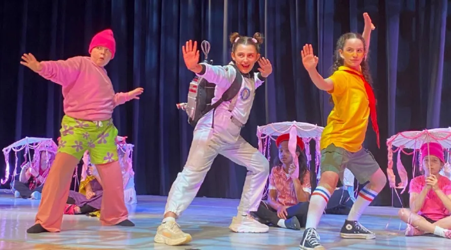 Three children performing a theatrical dance on stage, with two seated children holding pink parasols in the background.