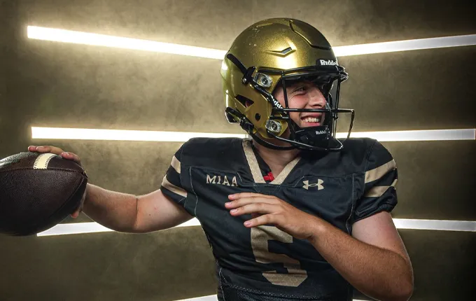 American football player wearing a gold helmet and black uniform poised to throw a football, smiling to the side.