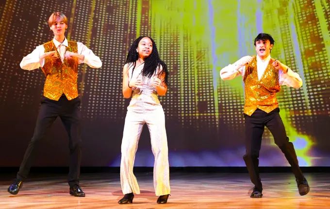 Three young performers dancing on stage with two wearing gold sequined vests and one in a shiny white jumpsuit.